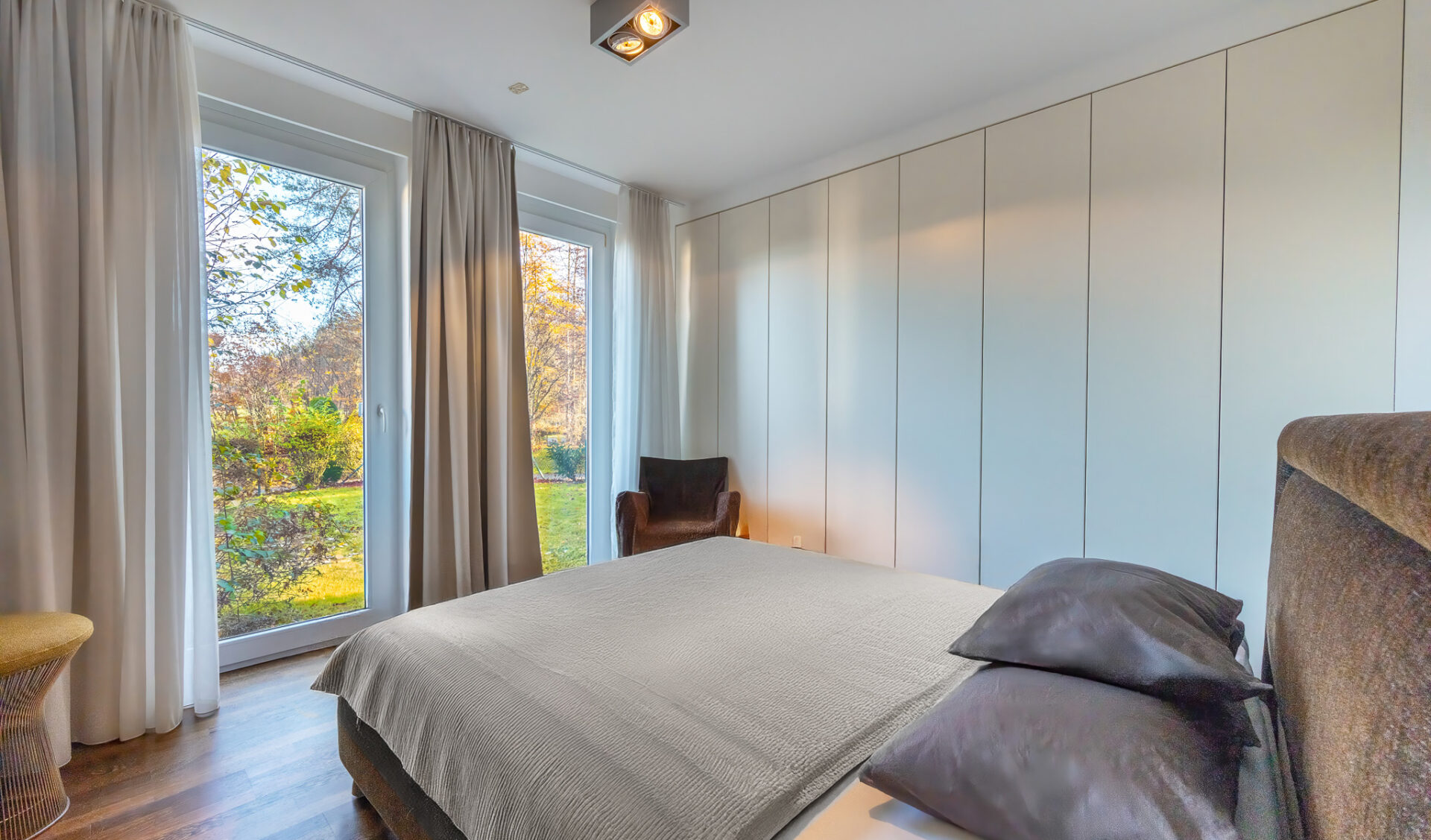 Helles Schlafzimmer mit großem Bett, bodentiefen Fenstern mit Blick in einen Garten, hellen Vorhängen und einer schlichten Einbauschrankwand.