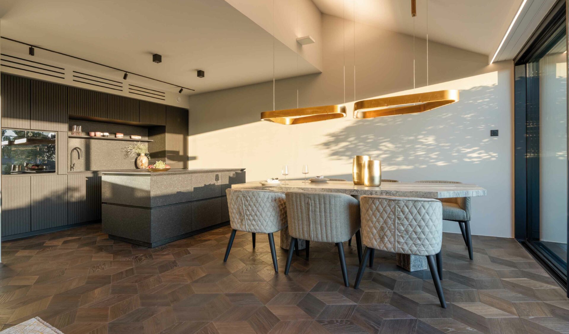 Offene Wohnküche mit Kücheninsel, Essbereich, goldenen Pendelleuchten und Fischgrätparkett.