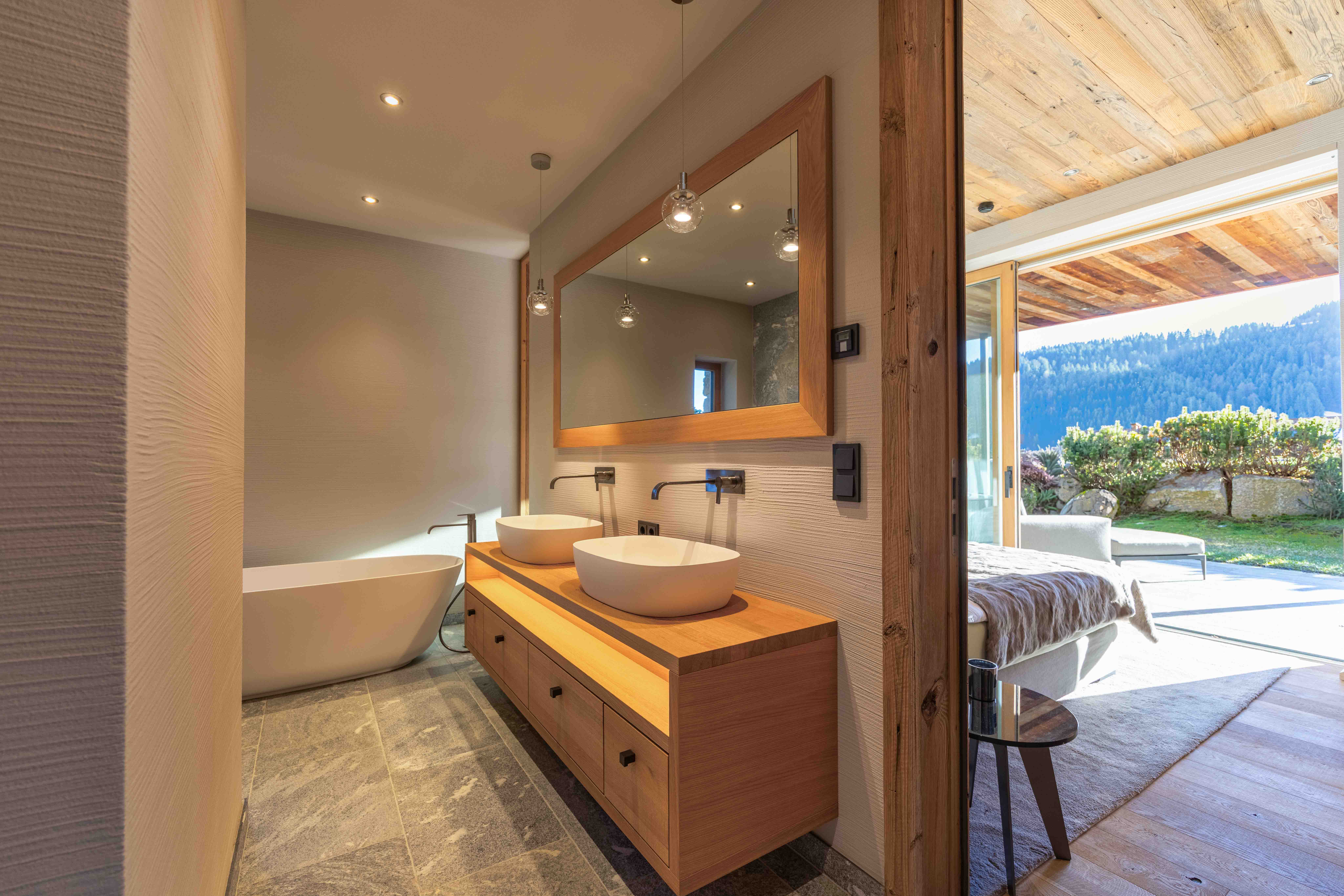 Modernes Luxuslandhaus-Badezimmer mit schwebender Holzablage, großem Wandspiegel und Durchgang zur Terrasse mit Alpenblick.