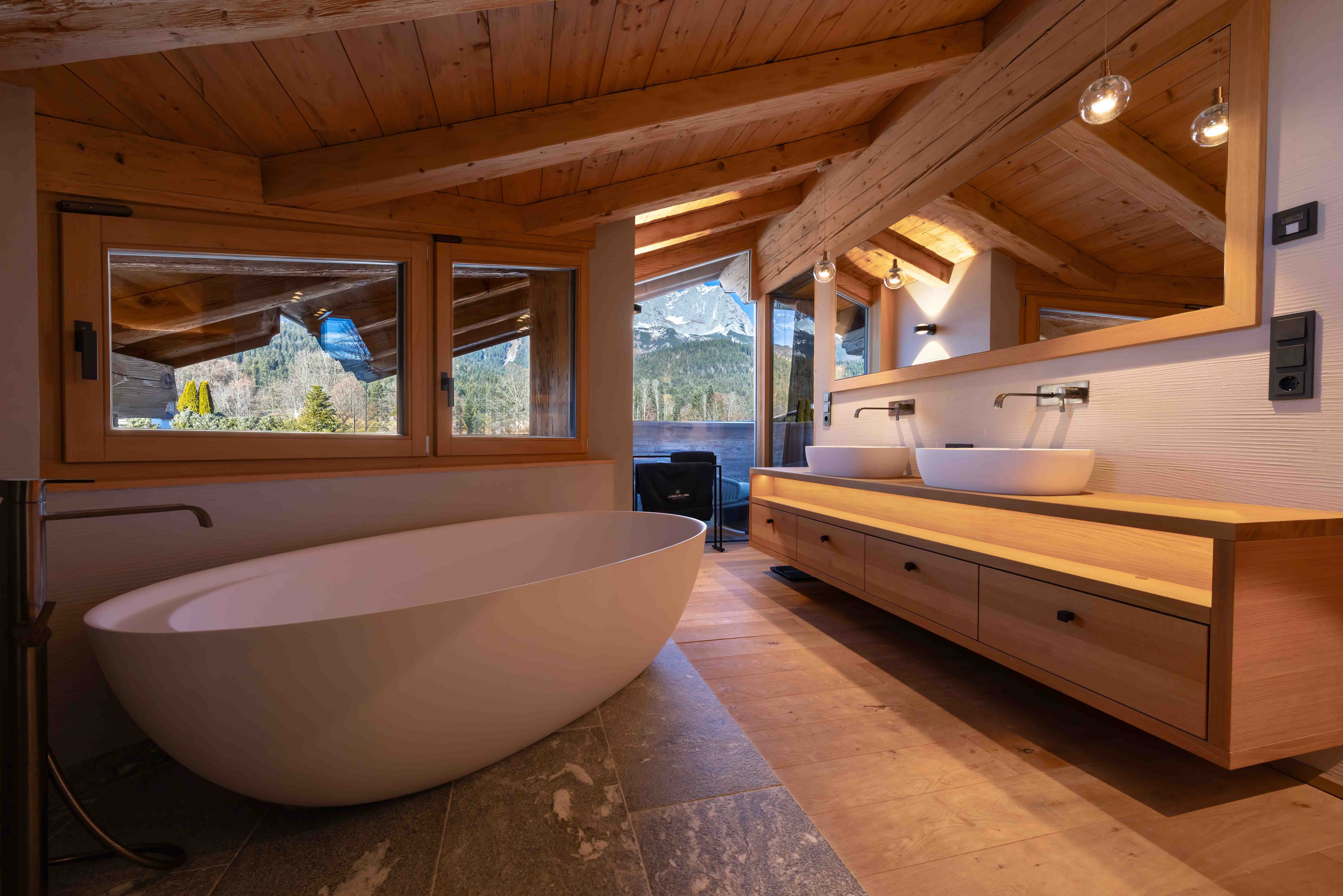 Modernes Chalet-Badezimmer mit Designer-Badewanne, Holz-Waschtisch, Naturstein-Boden und Panoramafenstern zu den Alpen.