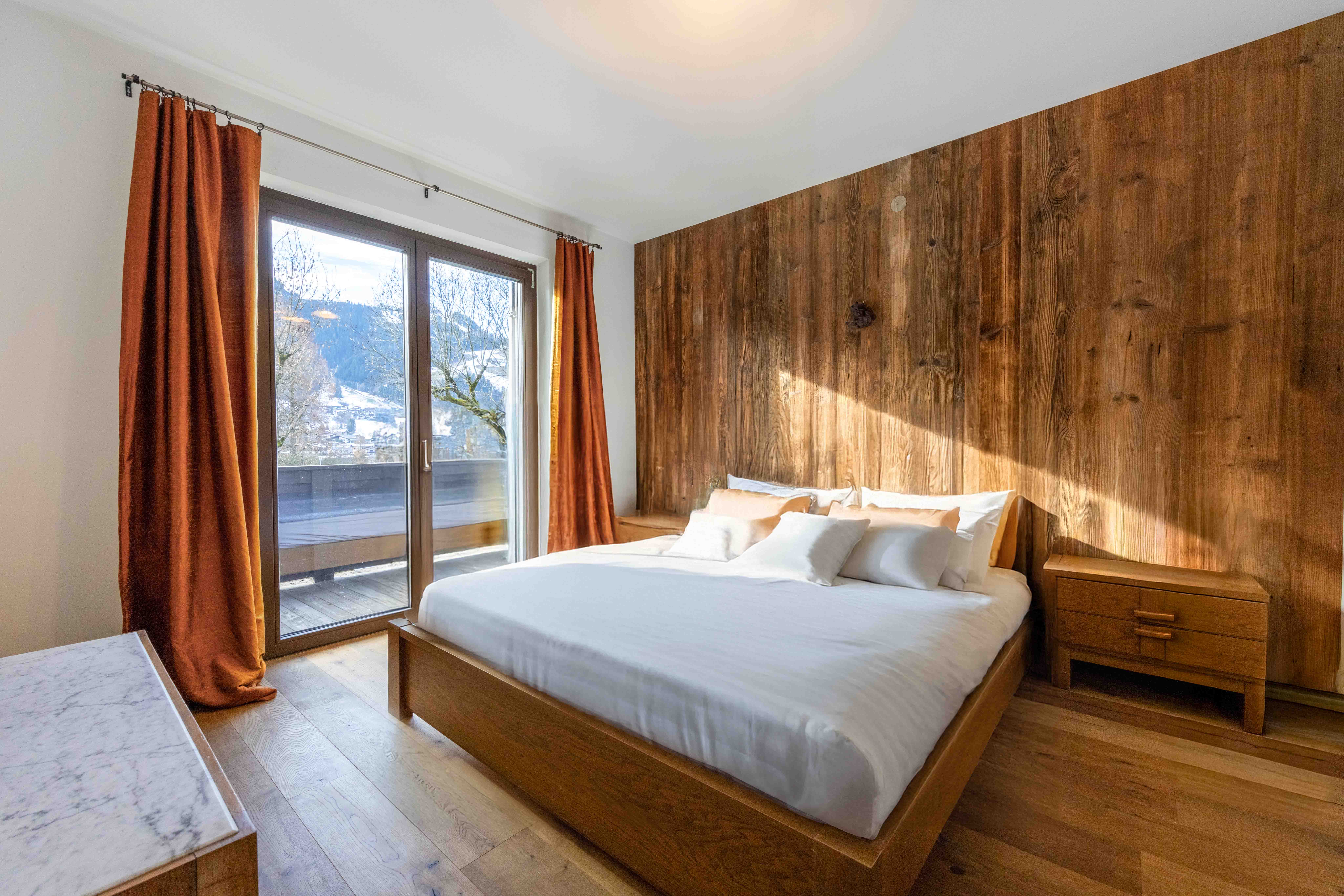 Helles Schlafzimmer im modernen Alpenstil mit Holzwand, Balkonzugang und Bergblick.