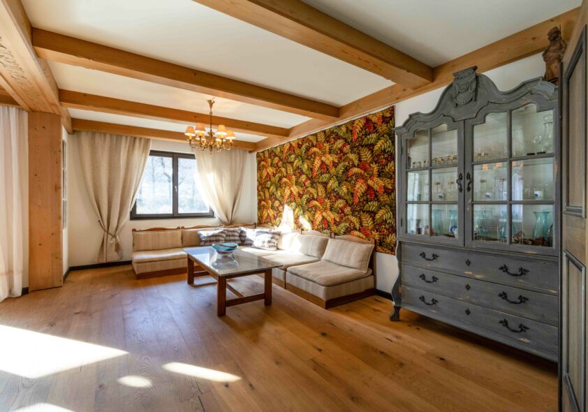 Gemütliches Wohnzimmer mit Holzbalkendecke, bunter Blumentapete und antikem Vitrinenschrank im Landhausstil