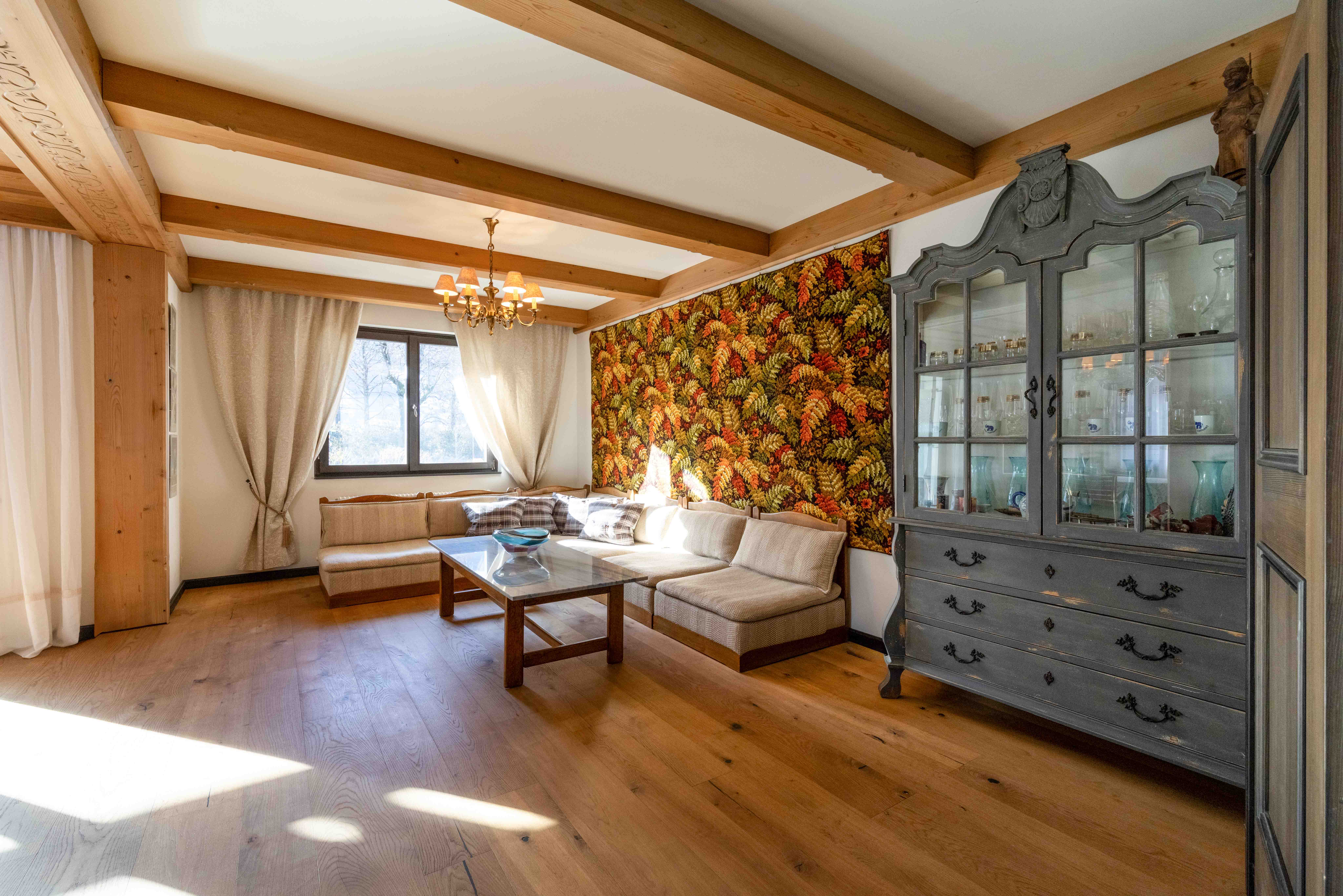 Gemütliches Wohnzimmer mit Holzbalkendecke, bunter Blumentapete und antikem Vitrinenschrank im Landhausstil