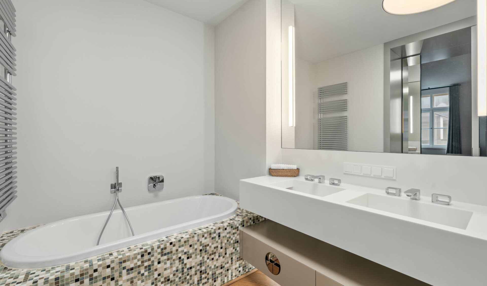 Luxusbadezimmer einer Neubauwohnung mit Badewanne in Mosaik-Optik, weißem Doppelwaschbecken und beleuchtetem Spiegelschrank.