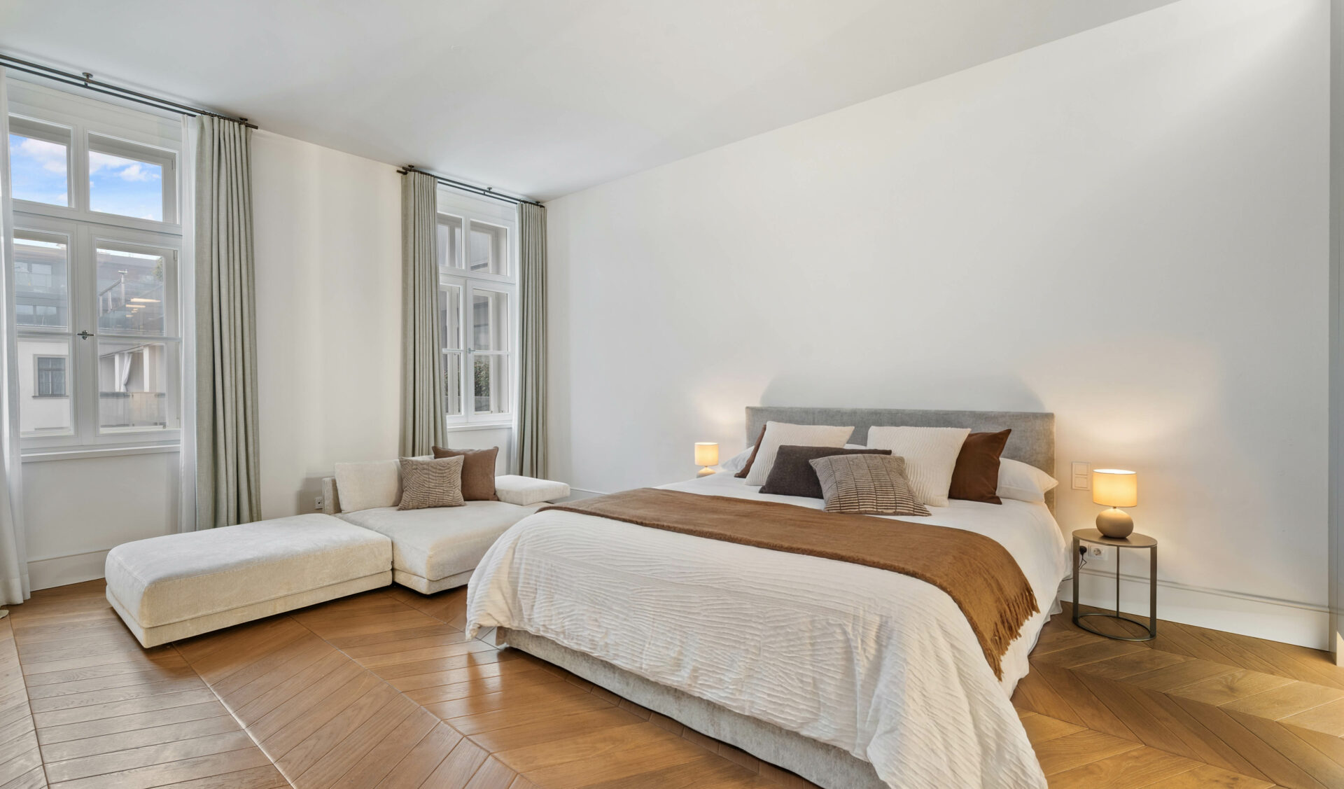 Großzügiges Schlafzimmer einer luxuriösen Altbauwohnung mit Kingsize-Bett, Chaiselongue, Fischgrätparkett und großen Fenstern.