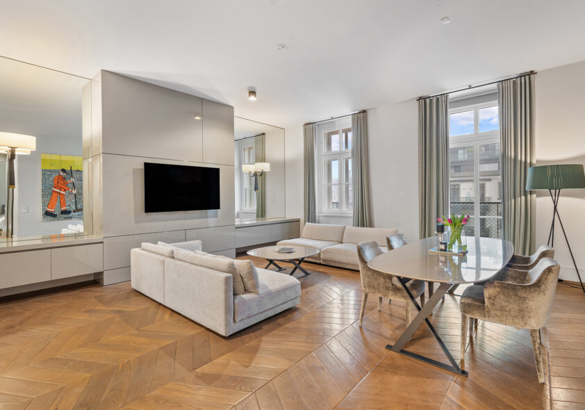 Modernes Wohnzimmer einer hellen Luxuswohnung mit hohen Decken, Fischgrätparkett, weißem Sofa, TV-Wand und Essbereich.