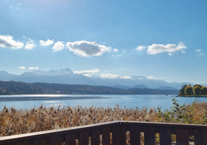 Panoramablick auf ruhigen Alpensee mit Schilf im Vordergrund und Holzterrasse.
