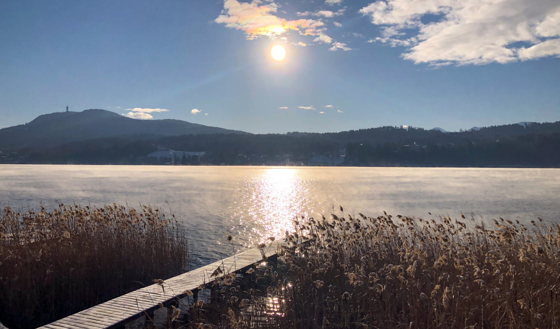 Idyllischer Seeblick mit Holzsteg und Bergpanorama bei Sonnenuntergang über ruhigem Gewässer mit Schilfufer.