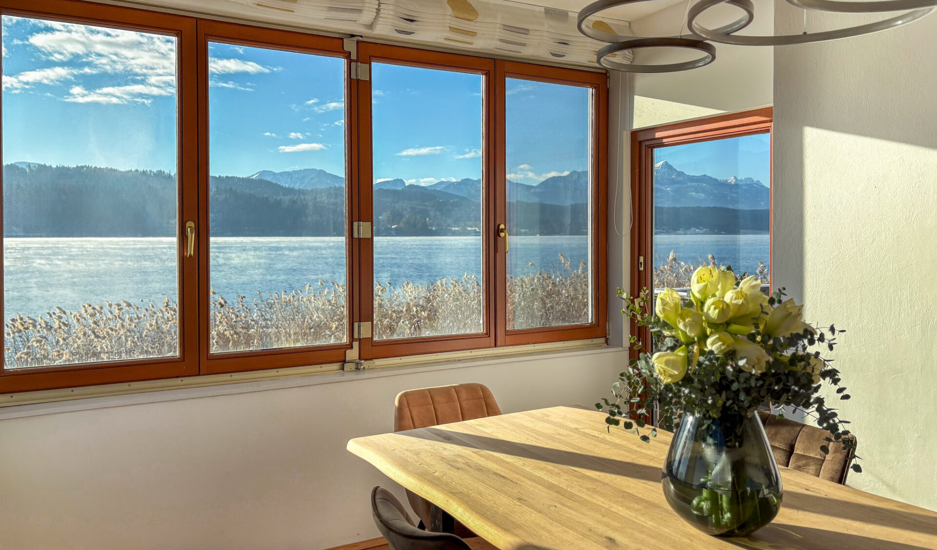 Lichtdurchflutetes Esszimmer mit Massivholztisch, Designer-Ringpendelleuchte und Panoramafensterfront mit See- und Bergblick.