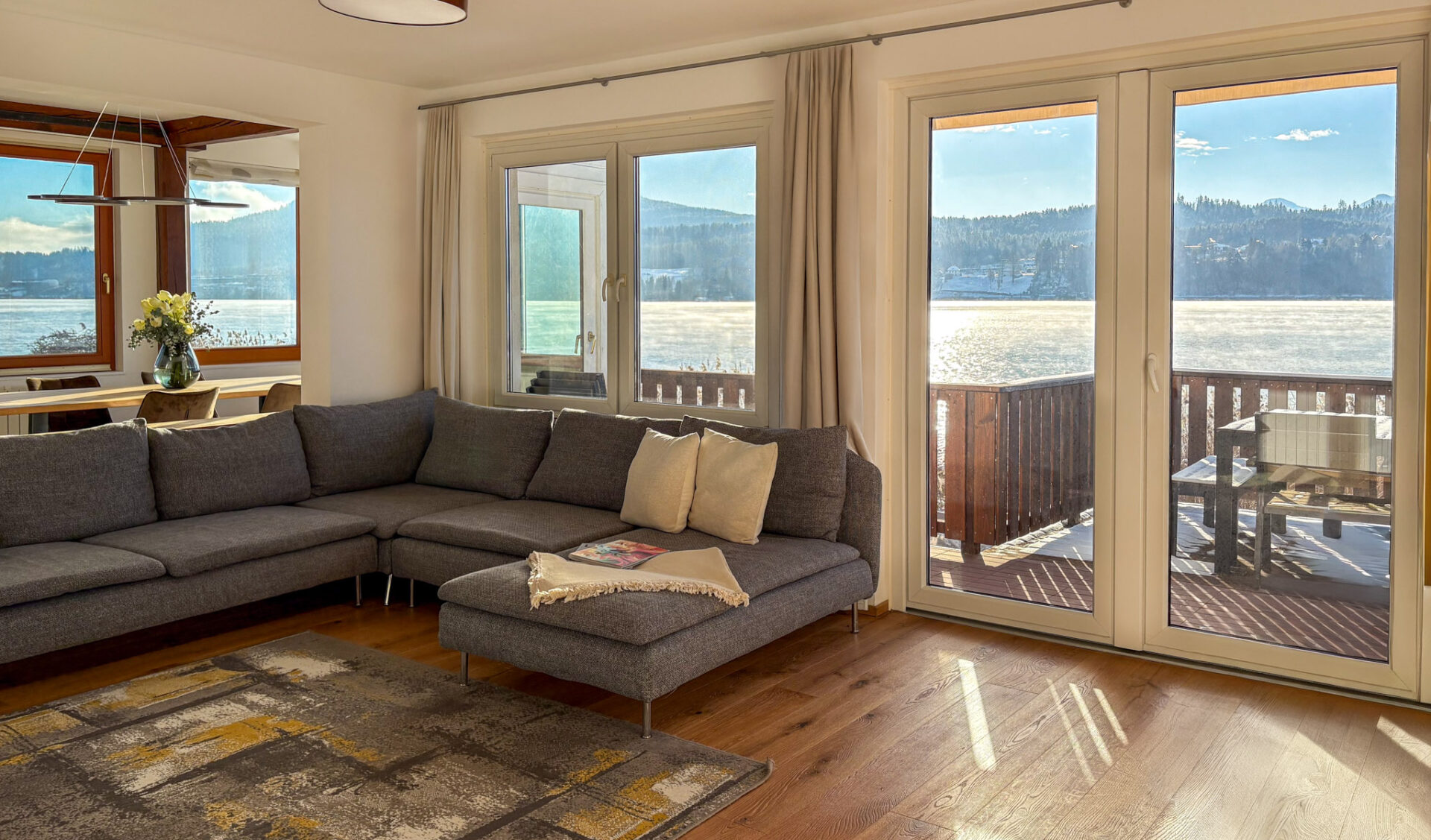 Geräumiges Wohnzimmer mit grauem Designer-Ecksofa, Parkettboden und bodentiefen Fenstern mit Panorama-Seeblick.