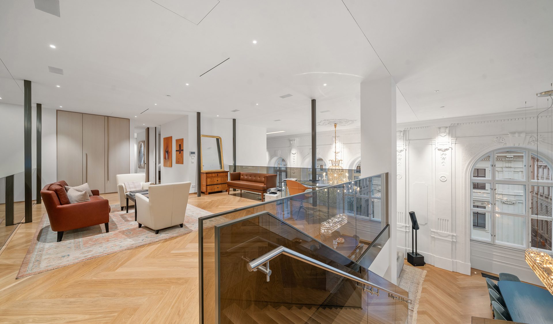 Großzügige Galerie-Lounge mit offenem Treppenabgang, Glasgeländer und Blick auf historische Stuckfassade.