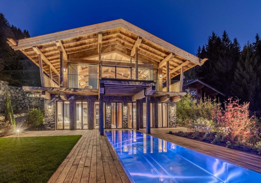 Premium-Bergchalet mit Infinity-Pool, LED-Beleuchtung, Naturstein und Panoramaverglasung bei Abendlicht.