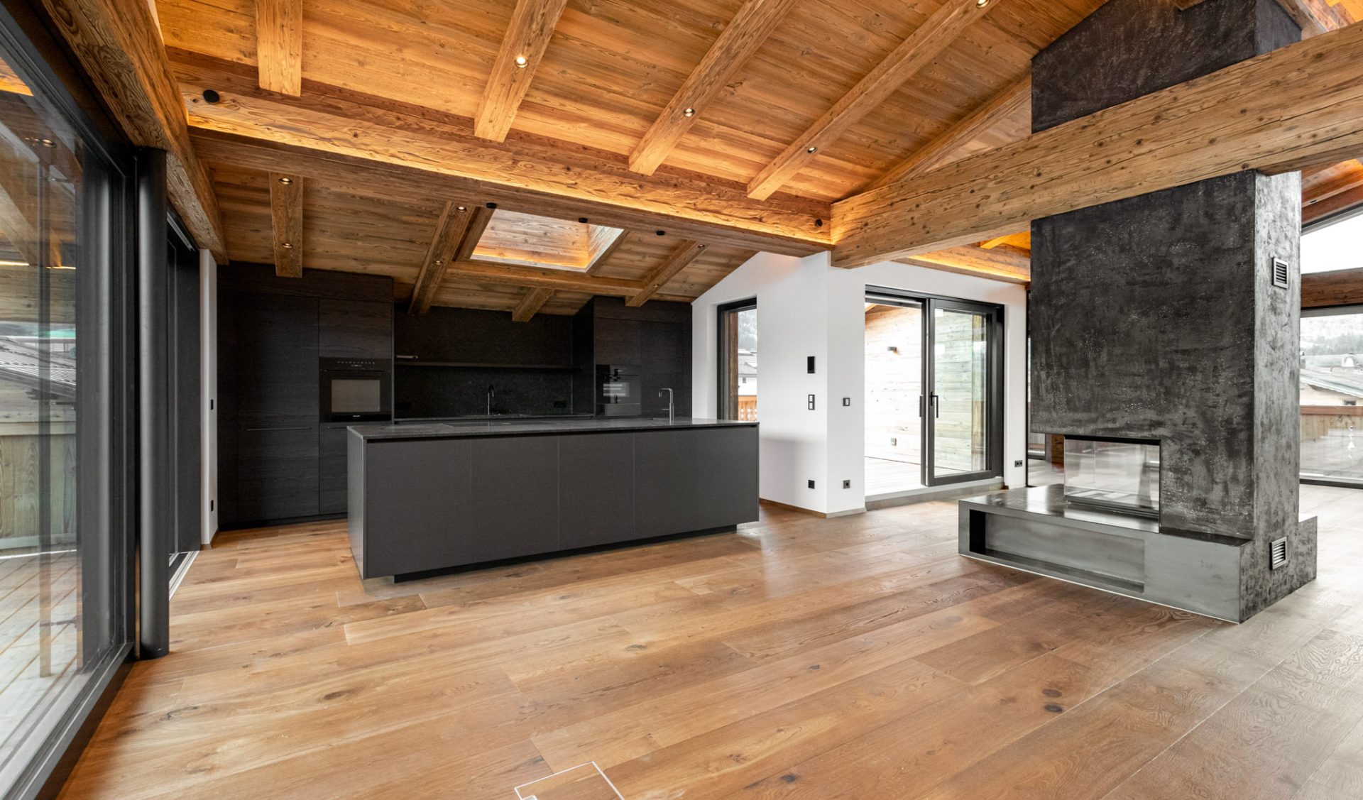 Offene Wohnküche mit schwarzer Kochinsel, Altholzbalkendecke, Kaminofen und Panoramaterrassenzugang.