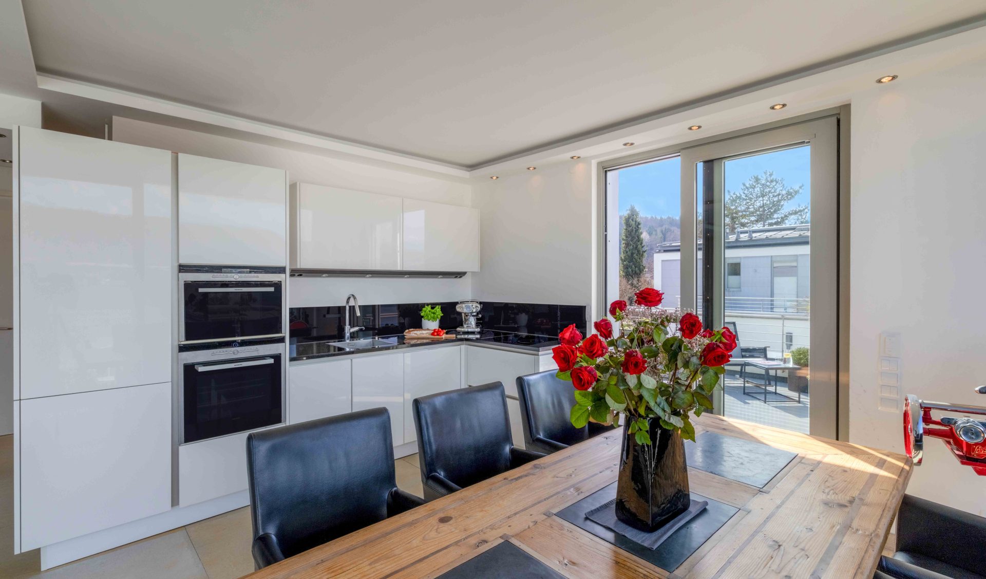 Exklusive weiße Hochglanz-Einbauküche mit Kochinsel, hochwertigen Geräten und angrenzendem Essplatz mit Zugang zur Terrasse.