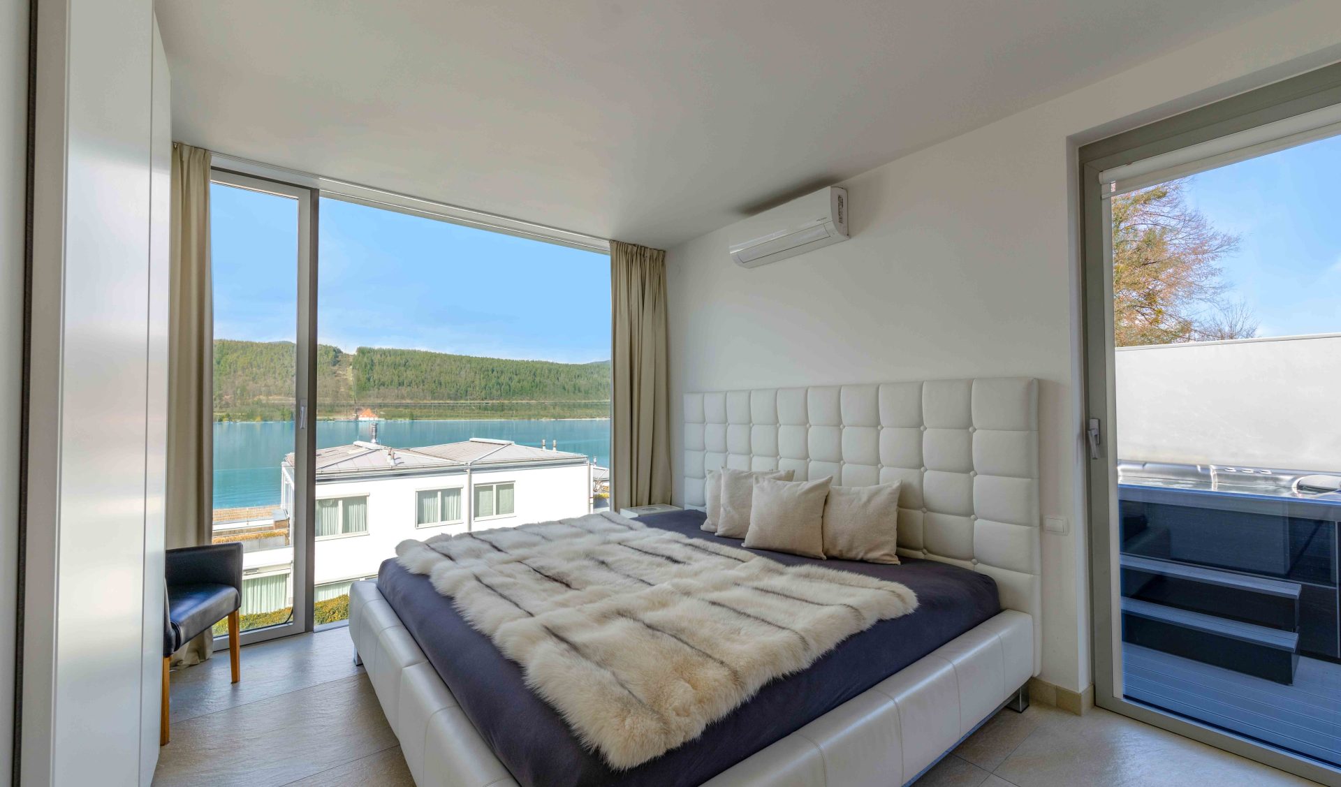 Luxuriöses Schlafzimmer des Wörthersee-Penthouses mit weißem Polsterbett, Felldecke und Panorama-Glasfront mit Blick auf das Wasser und die Berge.