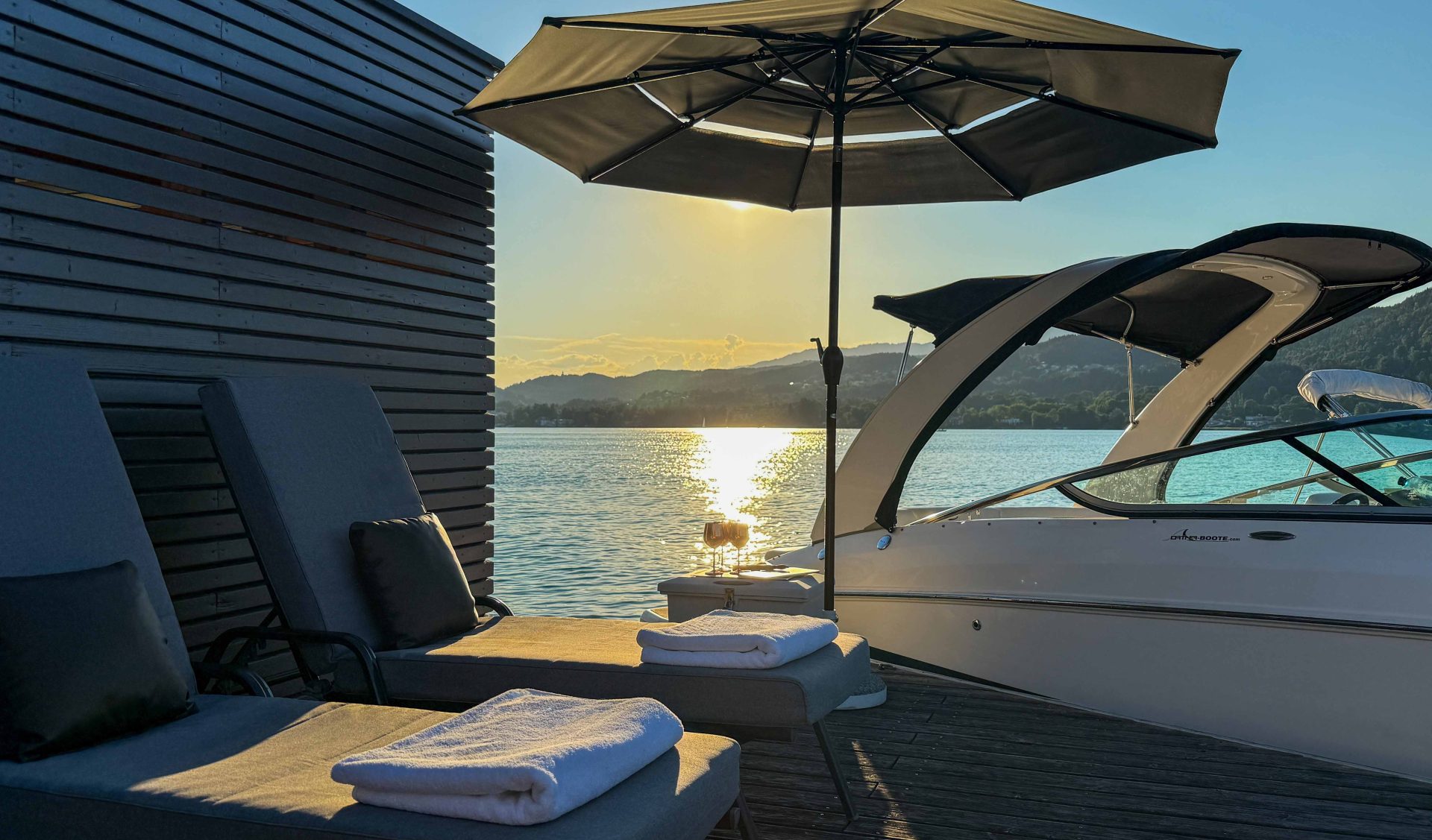 Privater Badesteg am Wörthersee bei Sonnenuntergang mit luxuriösen Sonnenliegen, Sonnenschirm und angelegtem Motorboot.