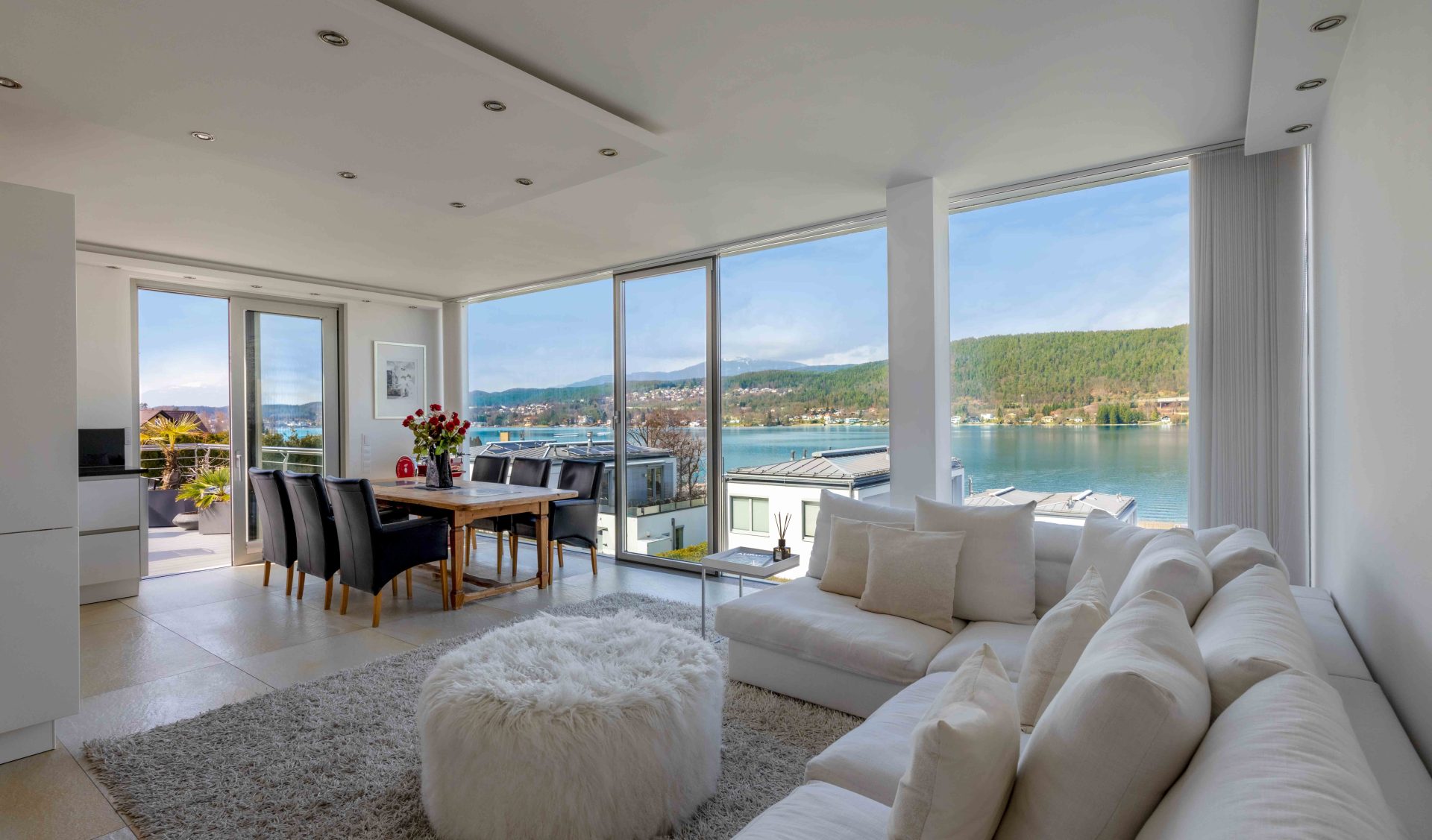 Lichtdurchfluteter Wohnbereich des Penthouses mit bodentiefen Fensterfronten, weißer Eckcouch und direktem Blick auf den Wörthersee.