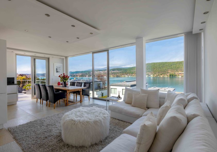 Lichtdurchfluteter Wohnbereich des Penthouses mit bodentiefen Fensterfronten, weißer Eckcouch und direktem Blick auf den Wörthersee.