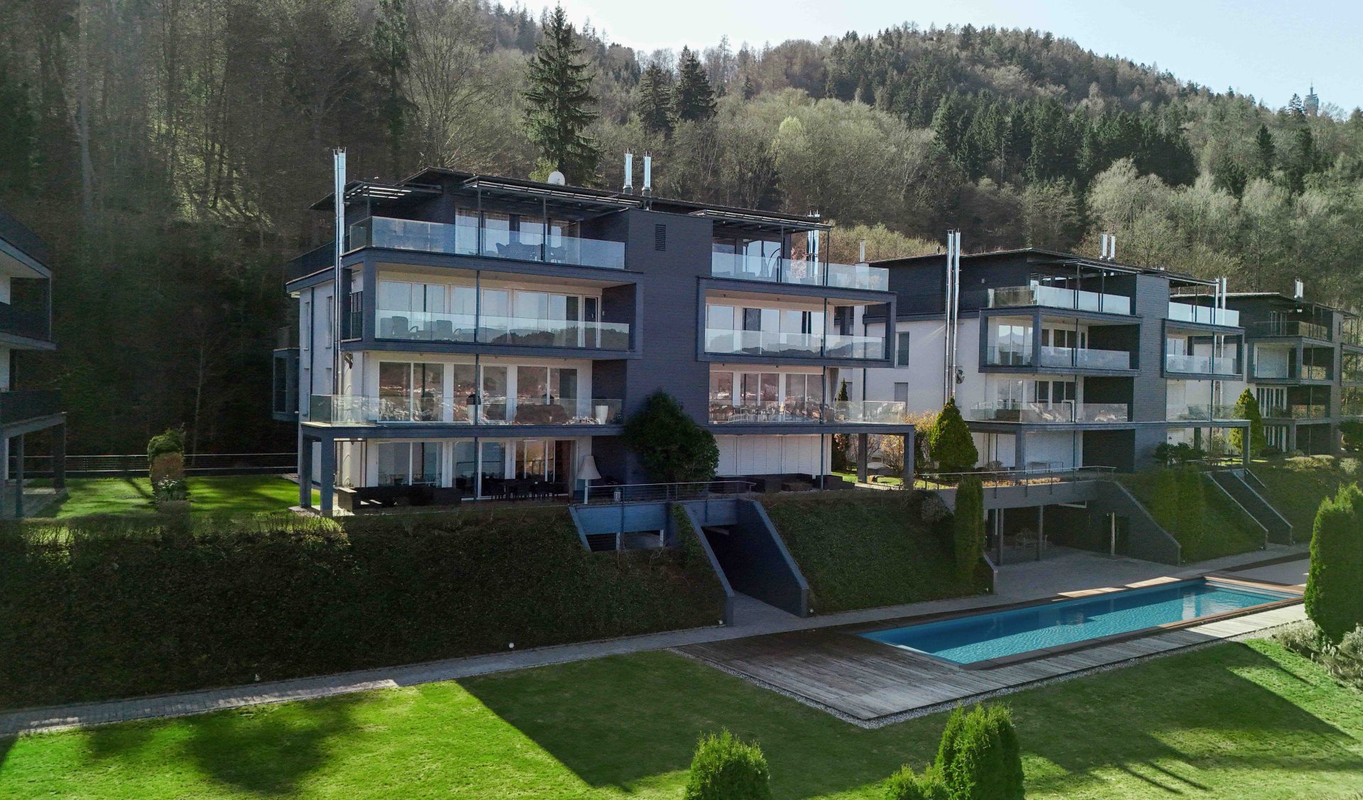 Moderne Terrassenwohnungen in Hanglage mit großen Glasfronten, Außenpool und gepflegter Gartenanlage vor einer bewaldeten Hügelkulisse.