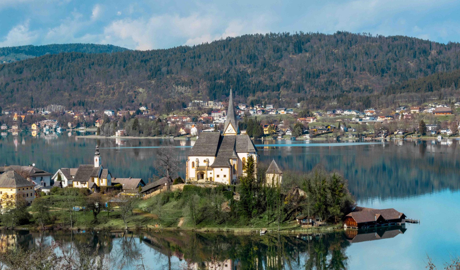 Idyllische Halbinsel Maria Wörth am Wörthersee mit der historischen Pfarrkirche vor bewaldeten Bergen in Kärnten.