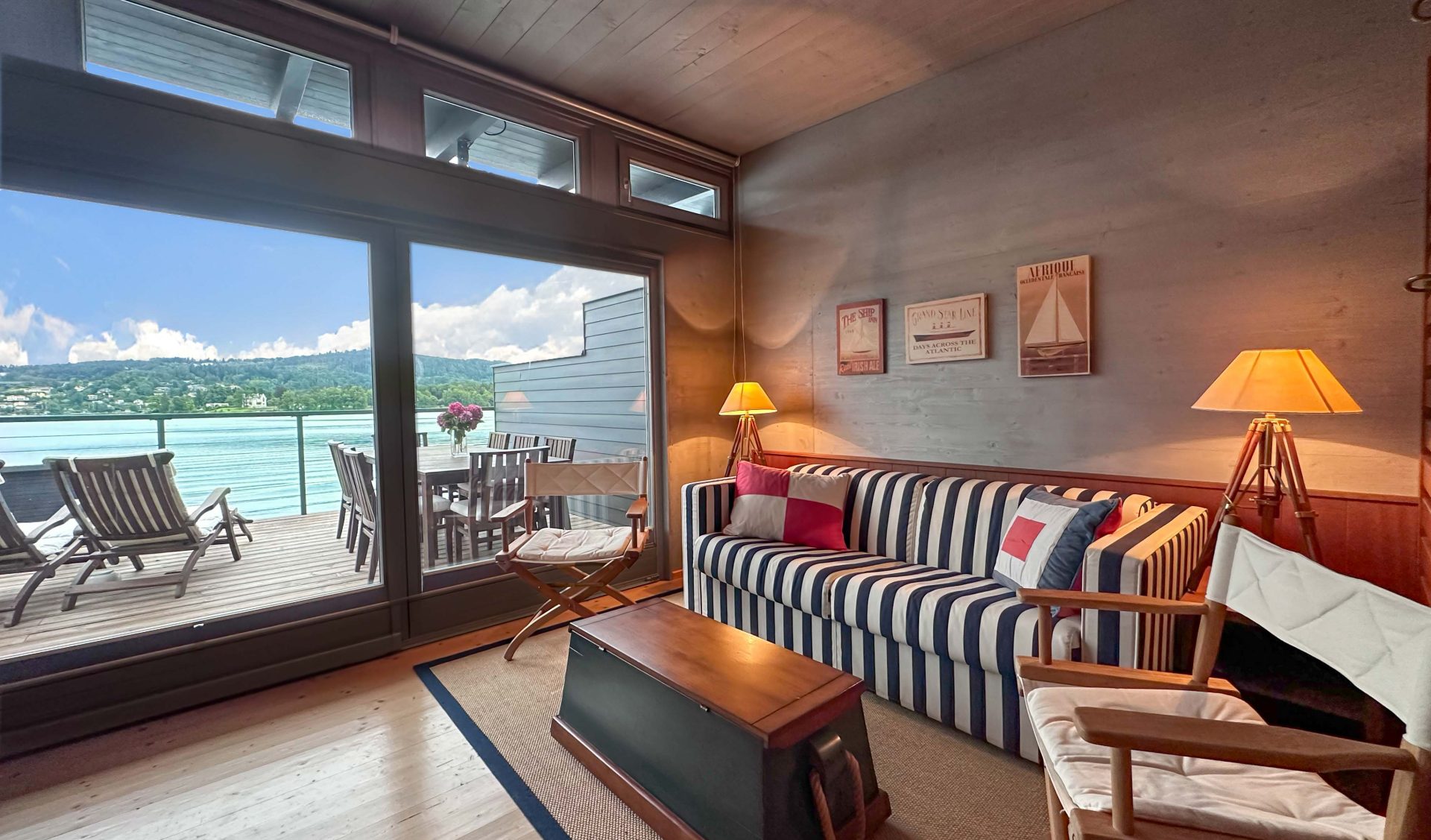 Bootshaus Obergeschoss mit gemütlichem Wohnraum im gestreiftem Sofa, Holztruhe als Couchtisch und großer Fensterfront mit direktem Blick auf den Wörthersee und die Terrasse.
