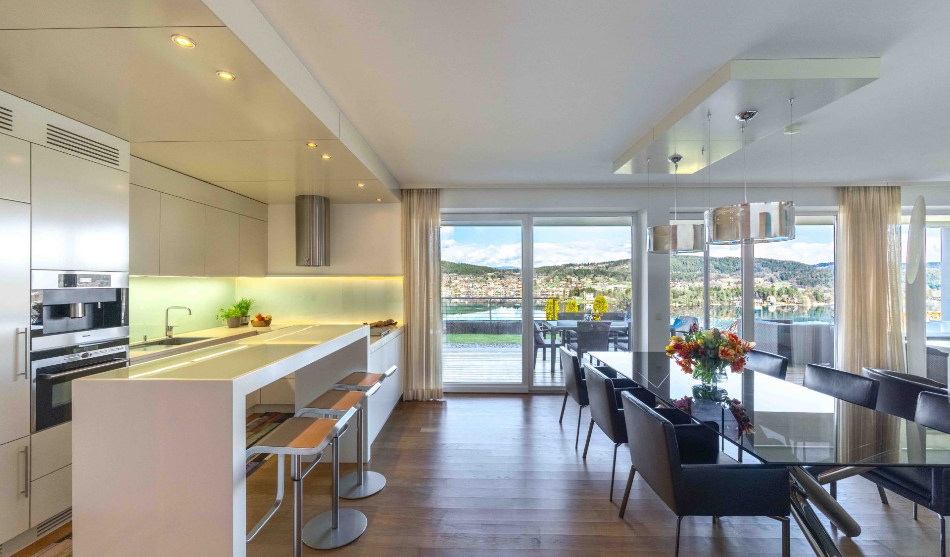Lichtdurchfluteter Wohnbereich mit Designer-Küche, Glastisch, Ledersesseln und Panoramablick auf den Wörthersee.