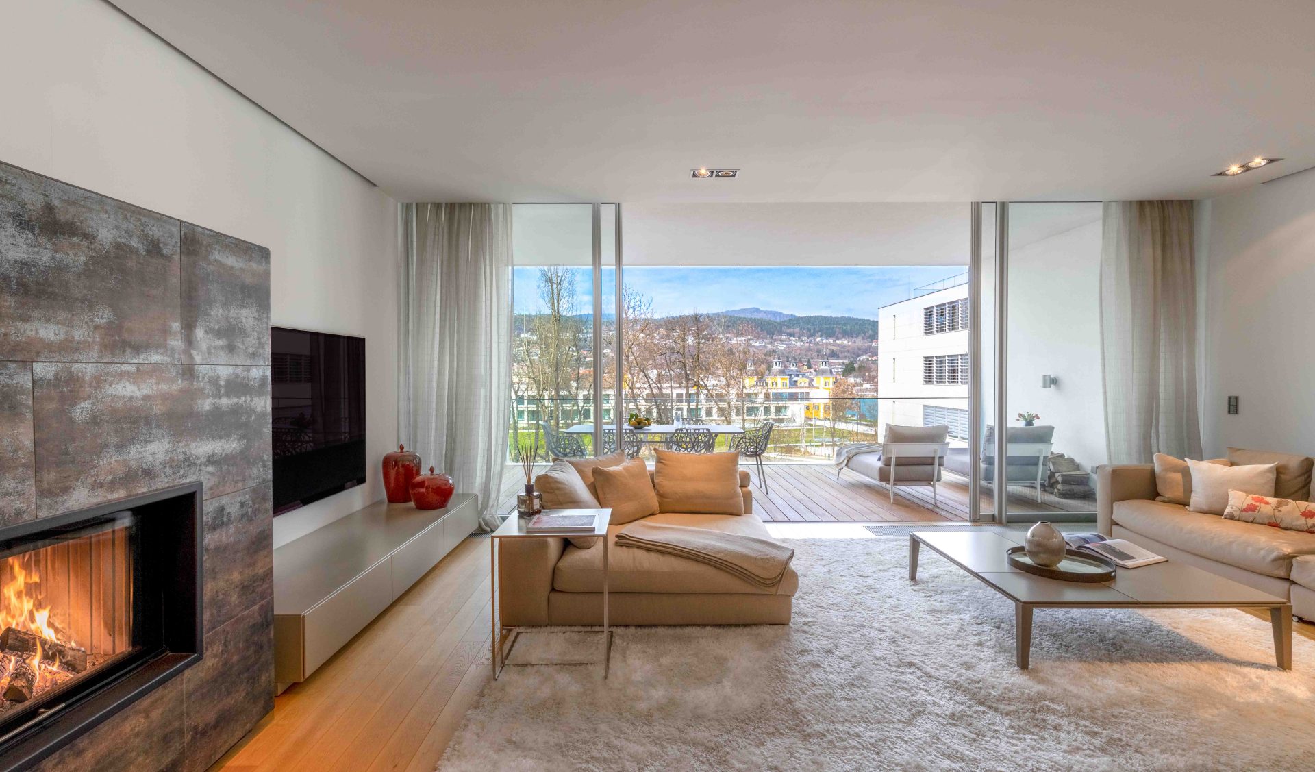 Offener Wohnbereich mit modernem Design-Kamin, heller Polstergruppe und großen Fensterfronten zur Seeseite im Schloss Velden.